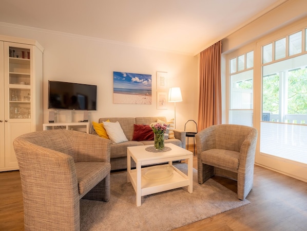 Un salon équipé d’un téléviseur à écran plat, d’une table basse, de deux fauteuils, d’un canapé, d’une lampe et d’une grande fenêtre avec des rideaux.