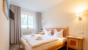 Una cama bien hecha con sábanas y toallas blancas, un cabecero de madera, una mesita de noche con lámpara y una ventana con cortinas.