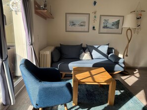 Ett vardagsrum med en soffa, två fåtöljer, ett soffbord och ett element. På väggen hänger två inramade tavlor och en väv av makramé. Det finns också ett fönster med gardiner.
