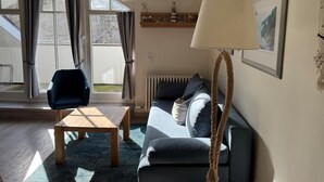 A cozy living room with a sofa, a coffee table, and a lamp. There is a window with curtains, a radiator, and a shelf with decorative items. - Comfort apartment with 1 bedroom and balcony on the top floor (Ostseebad Binz)