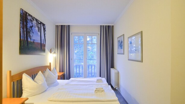 A hotel room with a double bed, a nightstand, a lamp, a window with curtains, and a painting on the wall. - Comfort apartment with 1 bedroom and balcony on the top floor (Ostseebad Binz)