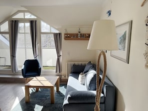 A living room with a sofa, a chair, a table, a lamp, a window with curtains, and a shelf with decorative items.