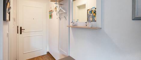 A hallway with a white door, a wooden bench, a mirror with a shelf, and a wall-mounted rack with items.