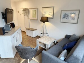 A modern living room with a grey sofa, a small white table, a grey armchair, a desk with a computer and speakers, a floor lamp, and framed artwork on the walls. - Comfort apartment with 1 bedroom and balcony on the top floor (Ostseebad Binz)