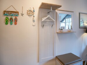 Une pièce avec une porte en bois, un banc blanc, une petite étagère ornée d’objets décoratifs et un panneau d’accueil avec des paires de tongs.