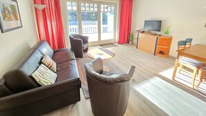 A living room with a sofa, armchair, and a small table. There is a TV on a wooden cabinet and a balcony with red curtains. - Comfort apartment with 1 bedroom and balcony on the upper floor (Ostseebad Binz)