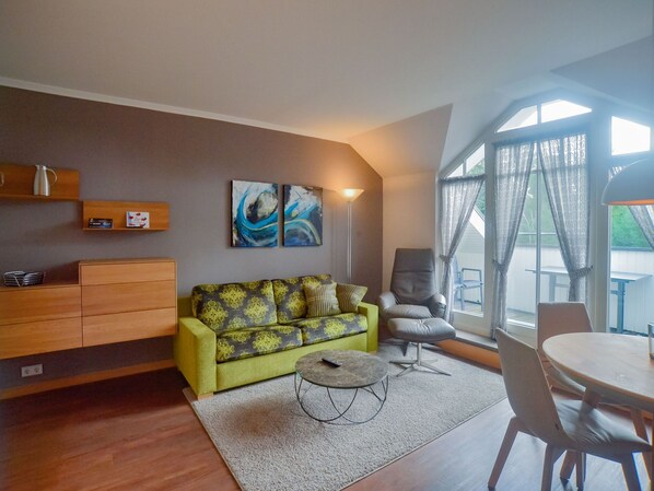 A modern living room with a green sofa, a round coffee table, and a dining area with a wooden table and chairs. - Comfort plus apartment with 1 bedroom and balcony on the top floor (Ostseebad Binz)