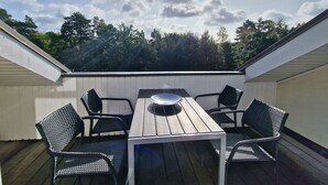 En utomhusveranda med ett träbord och fyra stolar, omgiven av ett vitt räcke, under en klar himmel med en synlig sol.