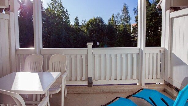 A covered outdoor area with a table and chairs, a blue lounge chair, and a view of trees and a building in the background. - Comfort apartment with 1 bedroom and balcony on the upper floor (Ostseebad Binz)