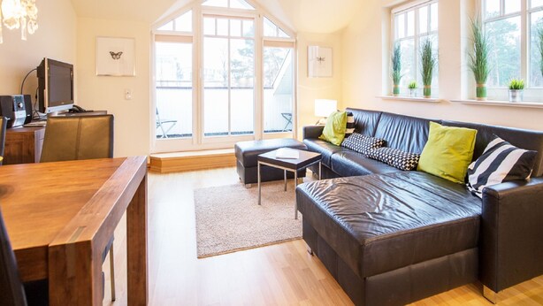 A modern living room with a leather sofa, wooden coffee table, and a desk with a computer. Large windows provide natural light and views of the outdoors. - Comfort plus apartment with 1 bedroom and balcony on the top floor (Ostseebad Binz)