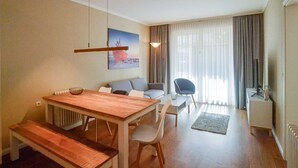 Una sala de estar moderna con una mesa de comedor de madera, un sofá y una silla. Hay un cuadro en la pared, una ventana con cortinas y un radiador.