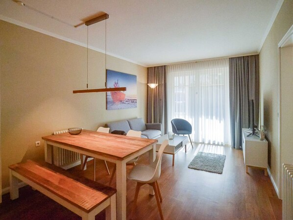 Ein modernes Wohnzimmer mit einem Holzesstisch, einer Couch und einem Sessel. An der Wand hängt ein Gemälde, ein Fenster mit Vorhängen und ein Heizkörper sind ebenfalls zu sehen.