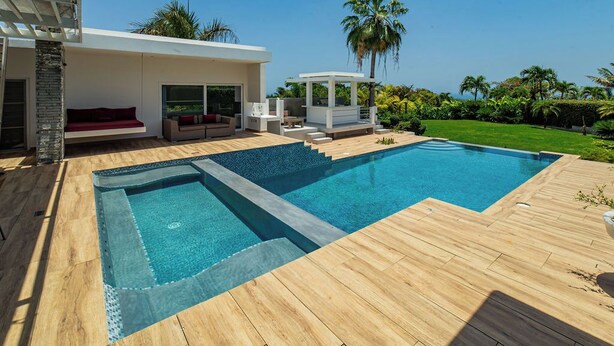 Een modern buitenzwembad met houten terras, een zithoek met rode bank en een overdekte constructie.