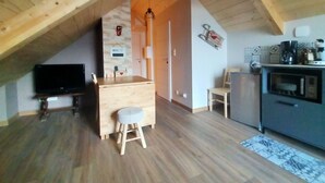 Een gezellige kamer onder het dak met houten balken, een televisie, een klein houten tafeltje met een krukje, een magnetron en een koffiezetapparaat.