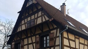 A traditional half-timbered building with a pitched roof, multiple windows with shutters, and a visible chimney. - GITE A LA PETITE FONTAINE-RDC  (Langensoultzbach)