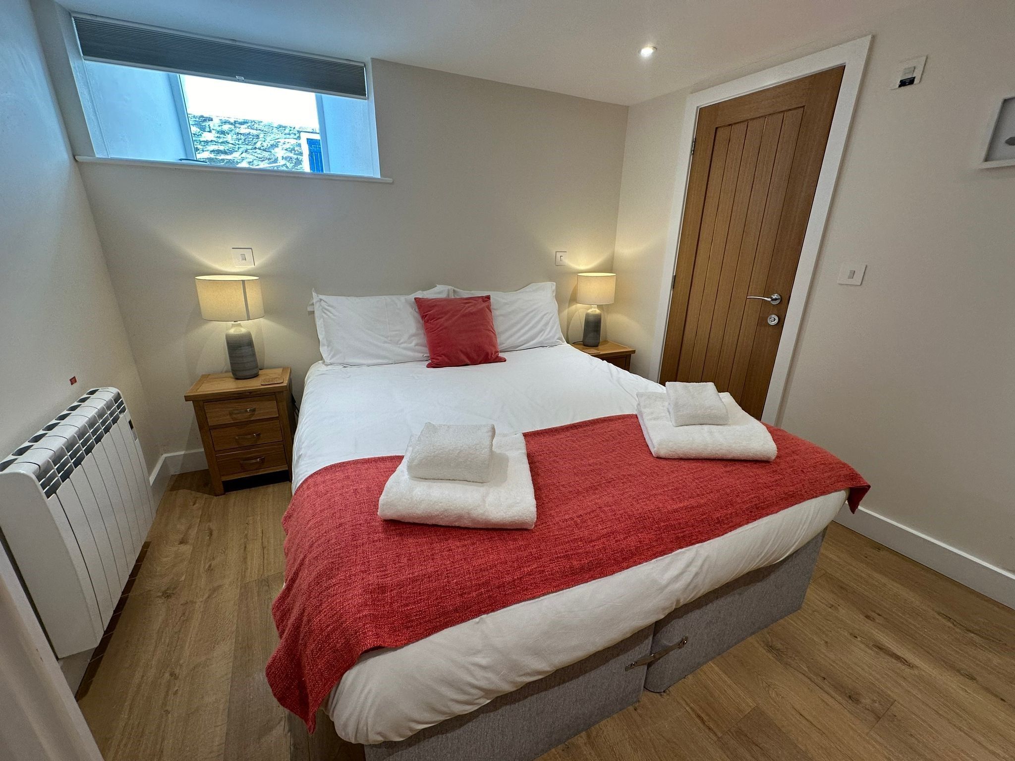 En snyggt bäddad säng med vita och röda sängkläder, två sänglampor, ett träbord vid sängen, ett element och ett fönster med utsikt över omgivningen.