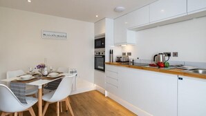 Ett modernt kök med vita skåp, en bänkskiva i trä och en matplats med vita stolar och ett bord dukat för två personer.