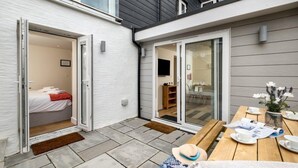 En modern uteplats med ett träbord dukat för middag, en skjutdörr i glas som leder in till ett sovrum och en liten trädgårdsdel med blommor.