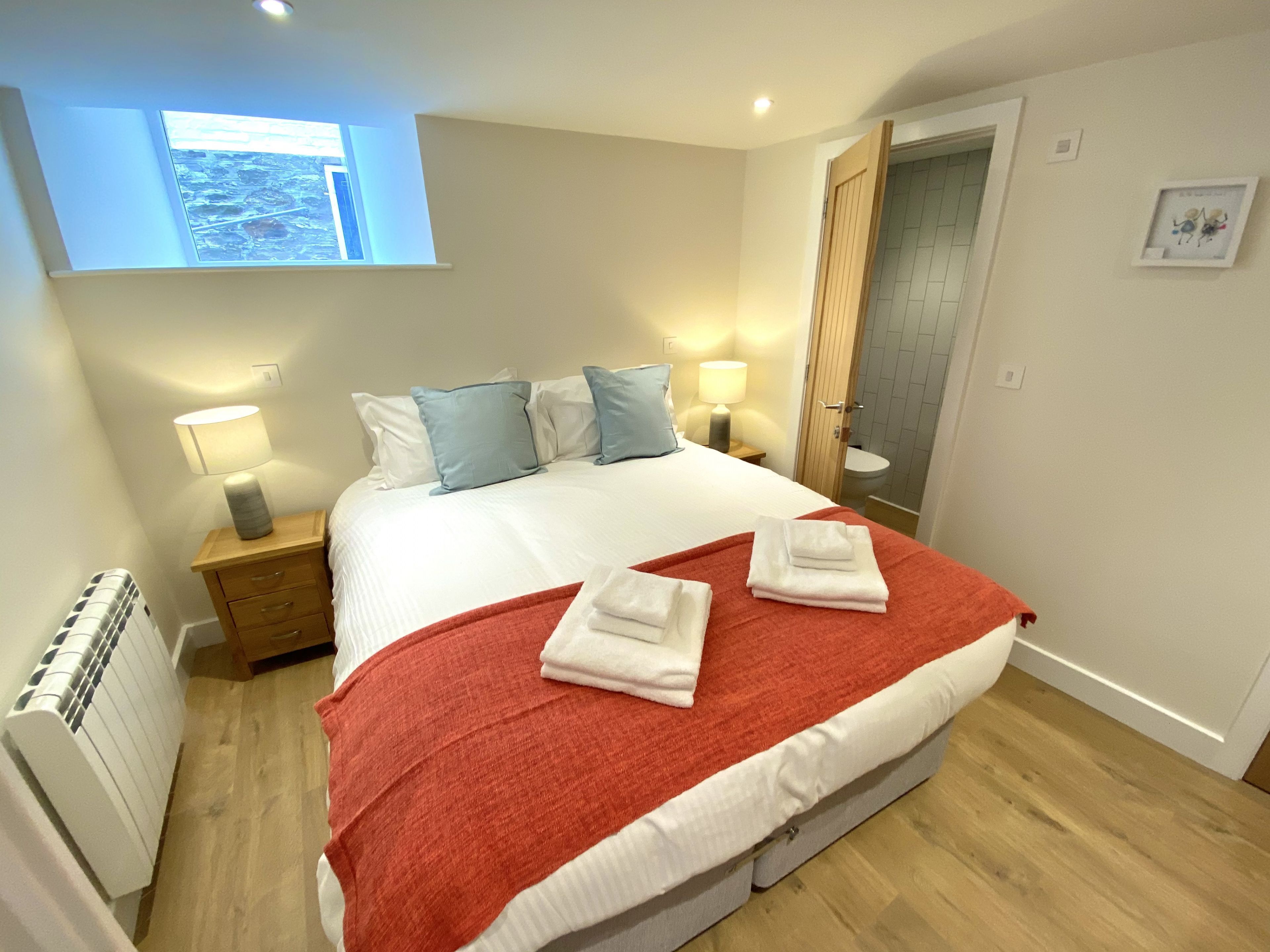 Ein ordentlich bezogenes Bett mit roter Decke und Kissen, eine Nachttischlampe und ein Fenster mit Blick.
