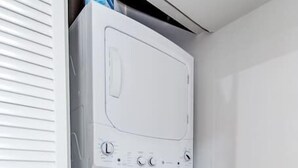Une machine à laver à chargement frontal, de couleur blanche, se trouve dans une buanderie dont le sol est en bois et le mur est blanc.