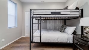 Une chambre avec un lit superposé, un plancher en bois, une porte blanche et une table de chevet avec une lampe.