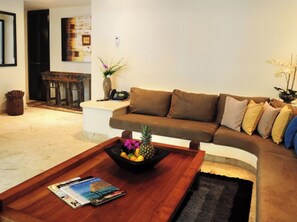 A living room with a brown sofa, a coffee table with magazines and fruit, and a vase with flowers.