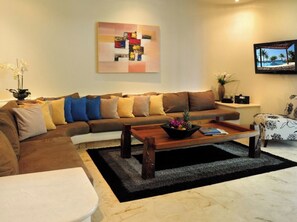 A living room with a sectional sofa, a coffee table with fruit, a painting on the wall, and a TV displaying a beach scene.