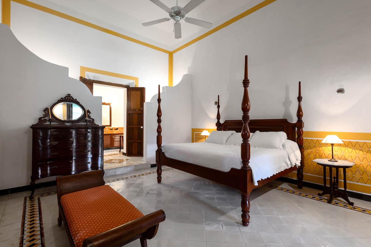 A four-poster bed with a wooden headboard, a bedside table with a lamp, a wooden dresser, and a bench.