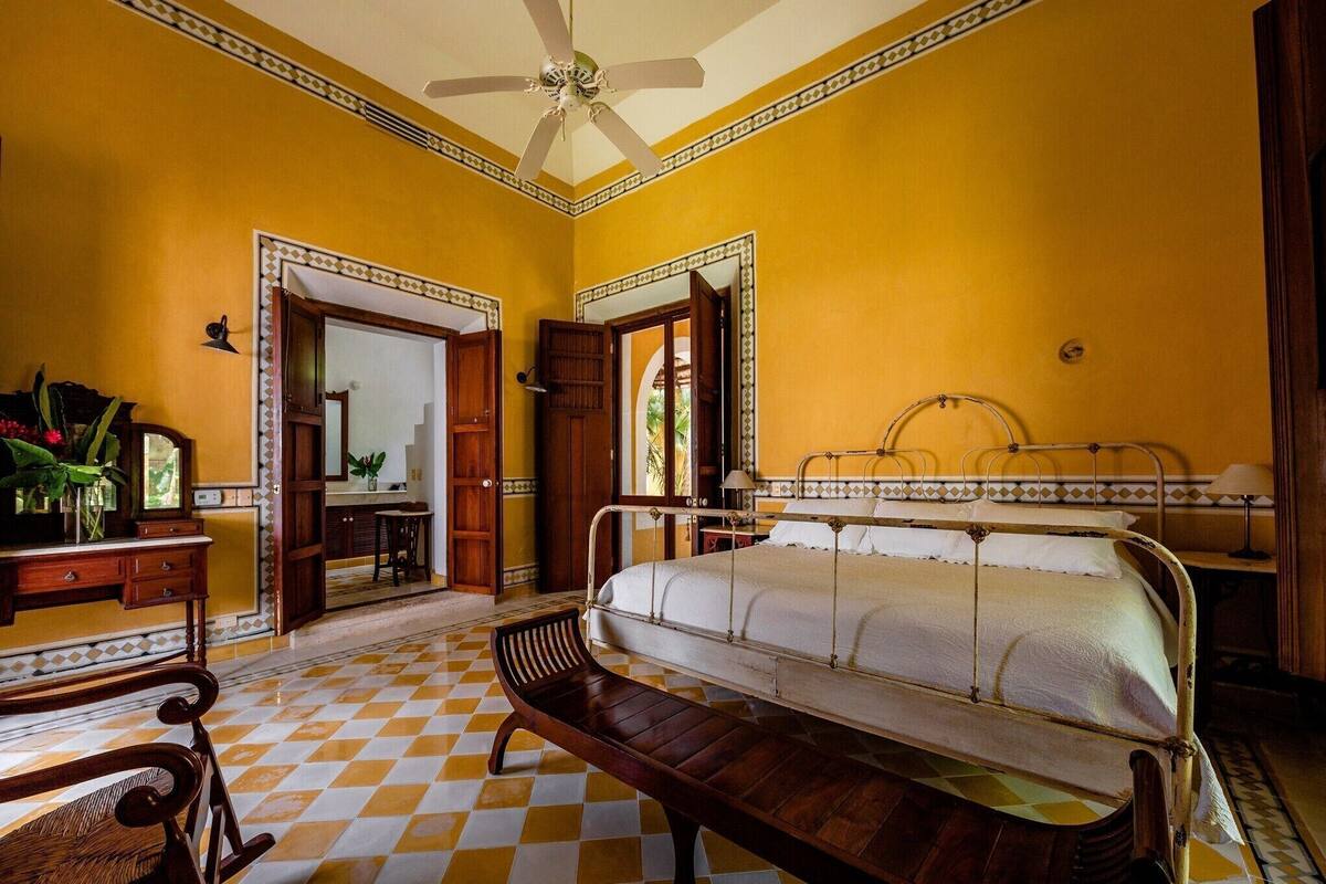 A room with a bed, a bench, a desk, and a ceiling fan.