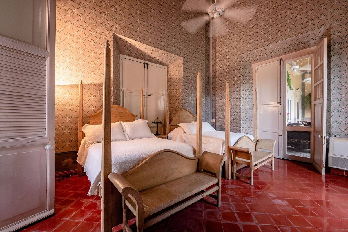 A four-poster bed with a wooden frame and white bedding, a wooden bench, and a red-tiled floor.