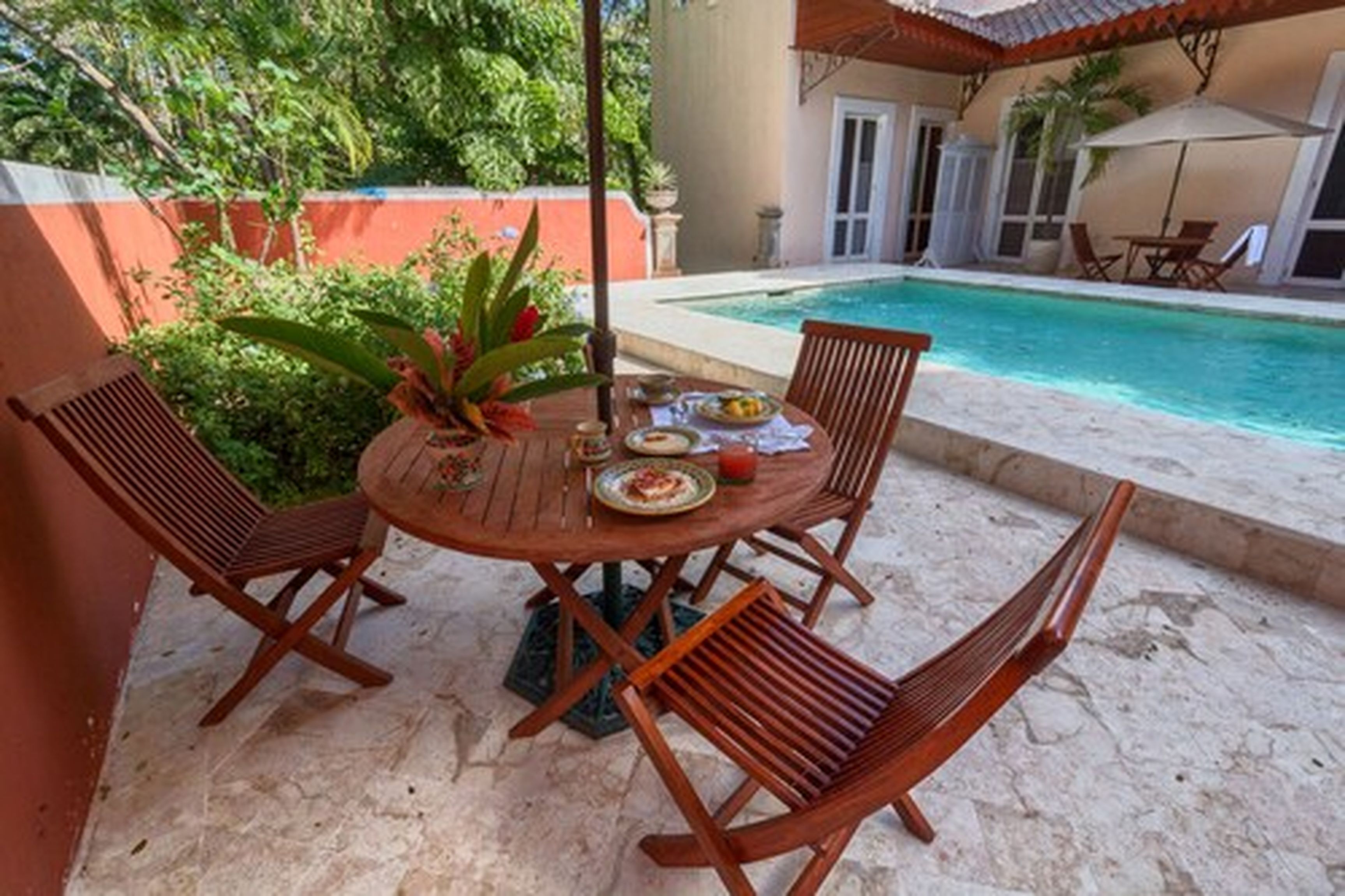Una mesa redonda de madera con cuatro sillas, una pequeña planta en maceta y un plato de comida en un patio con una piscina al fondo.