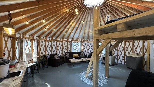 Une yourte dotée d’un plafond en bois, d’un luminaire central, d’un canapé, d’une table basse et d’un coin cuisine avec un poêle.