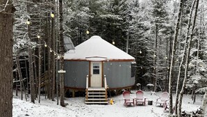 Een besneeuwde hut met een houten trap die naar een deur leidt, omringd door hoge bomen.