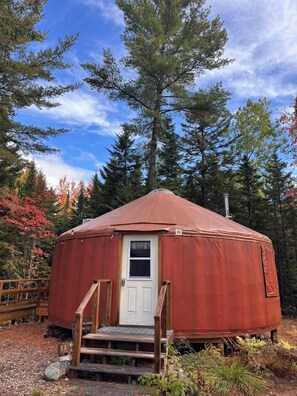 A round, red yurt with a white door and wooden steps leading up to it, surrounded by trees and a wooden deck.