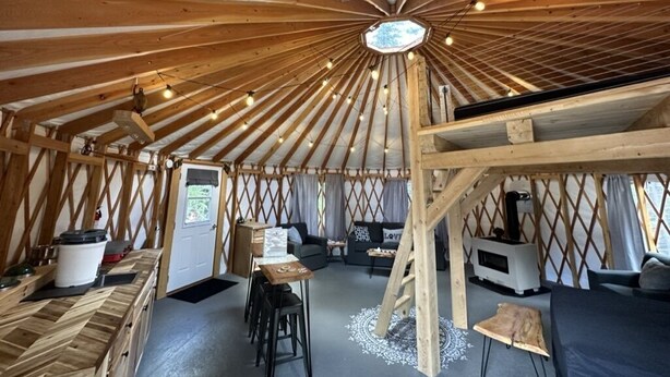 A yurt interior with a central wooden beam structure, a sofa, a small table, and a television. - The Swift Glamping Yurt in Western Maine (Bethel)