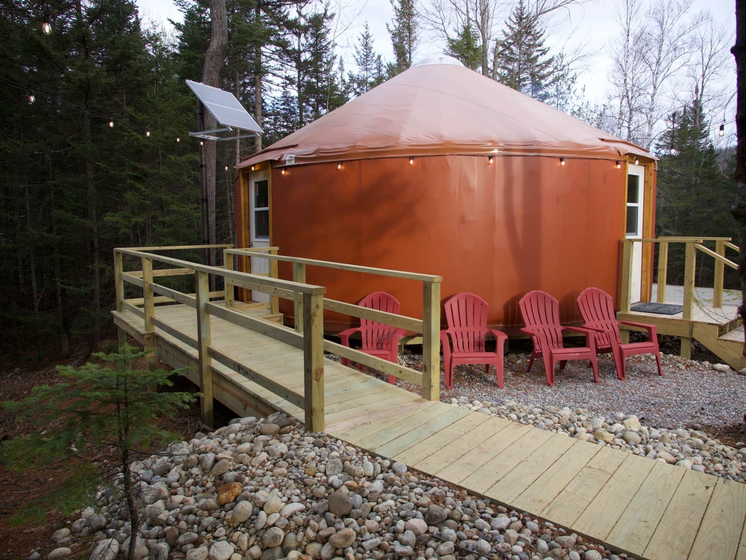 A wooden deck leading to a large, orange-roofed structure with a solar panel on the roof. There are five red chairs arranged in front of the structure.