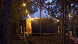 Une grande structure en forme de tente, avec un toit en dôme, située dans une forêt, entourée d’arbres, et dotée d’une terrasse en bois menant à l’entrée.