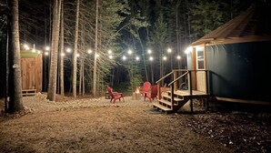 Una cabaña rústica con terraza de madera y zona de estar al aire libre, rodeada de árboles altos y luces colgantes.