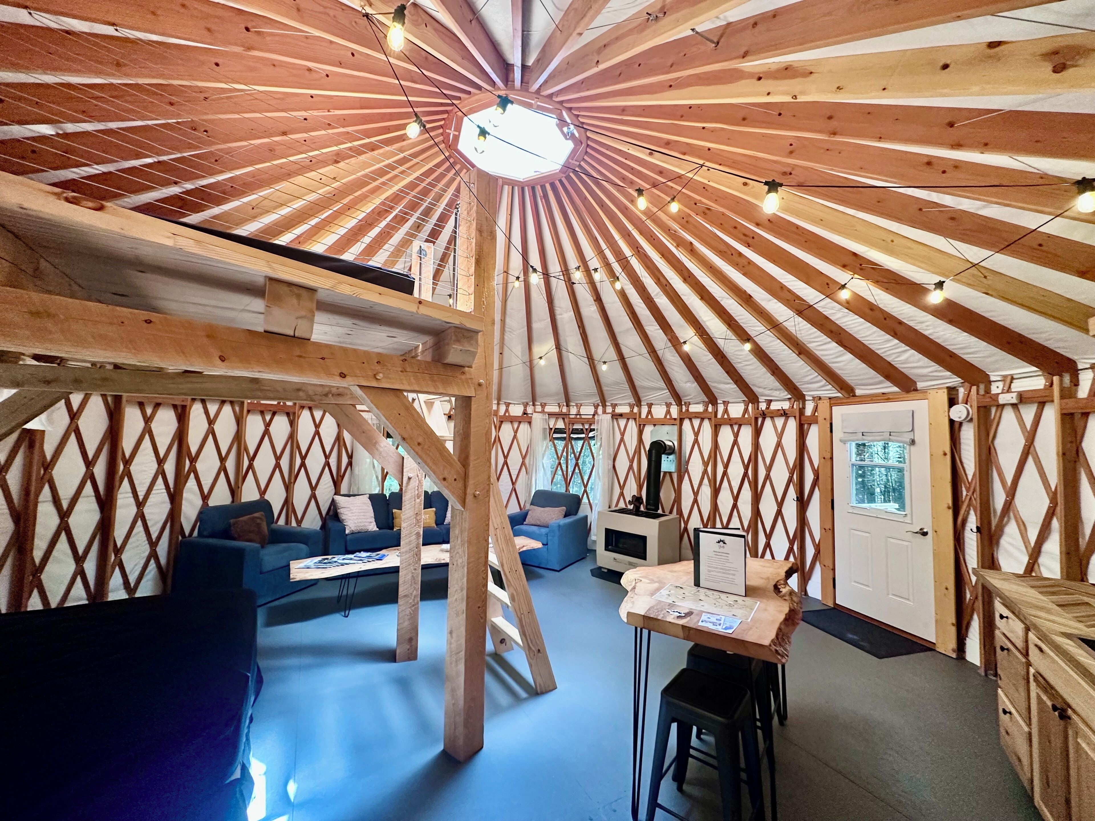 Interieur van een joert met houten balken, een centrale lamp, een bank, een tafel en een deur.