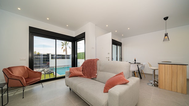 A modern living room with a sofa, a coffee table, and a dining area with chairs. There is a large window providing a view of the outdoors. - Villa Camilla Roda Golf (Roda)