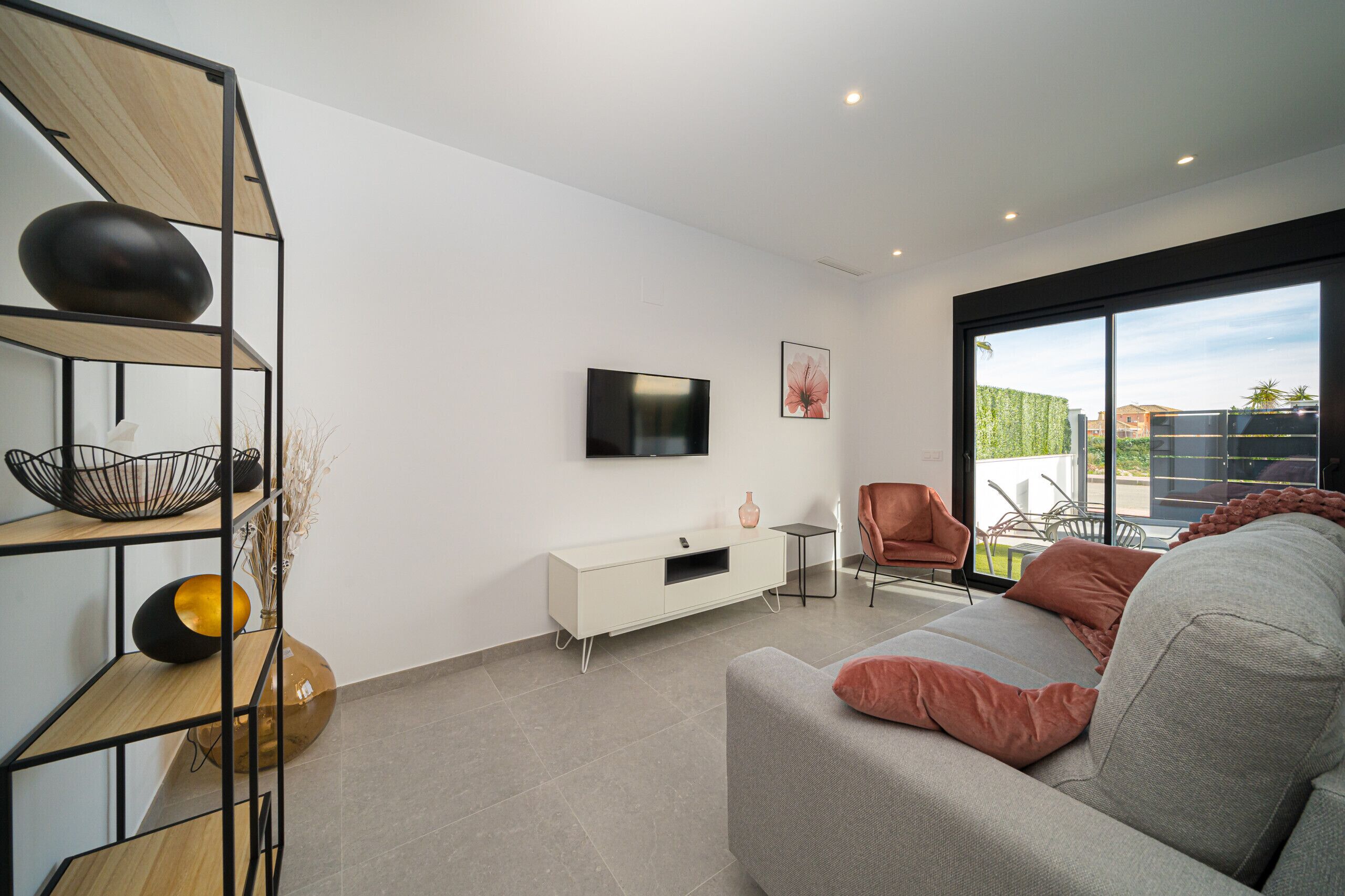 Ein modernes Wohnzimmer mit einem grauen Sofa, einer weißen TV-Konsole und einem großen Fenster mit Blick ins Freie.