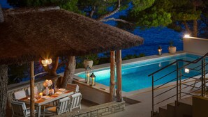 Zona de piscina exterior con área de comedor bajo un techo de paja, rodeada de árboles y con vistas a un cuerpo de agua.