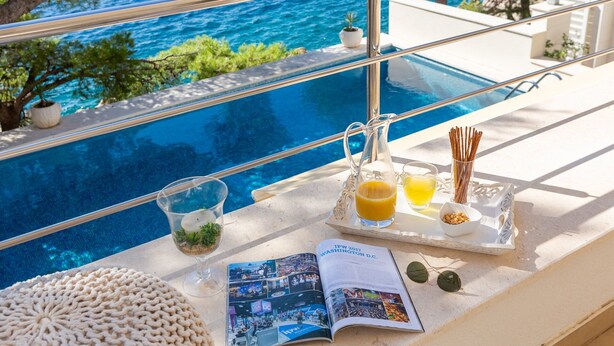 Un ambiente junto a la piscina con vistas despejadas al mar, una jarra de cristal con jugo de naranja, un vaso de bebida amarilla, una revista y una bandeja con aperitivos.