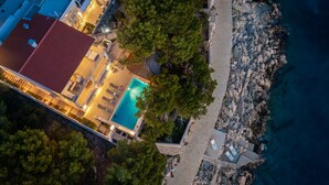 Vista aérea de una propiedad costera con piscina, un edificio de techo rojo y un litoral rocoso.