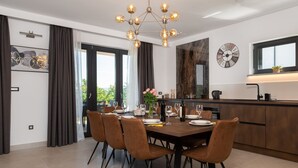 A modern dining area with a wooden table set for six, surrounded by brown upholstered chairs. A decorative chandelier hangs above, and a large clock is mounted on the wall. - Luxury Villa Phoenix with Pool (Labin)