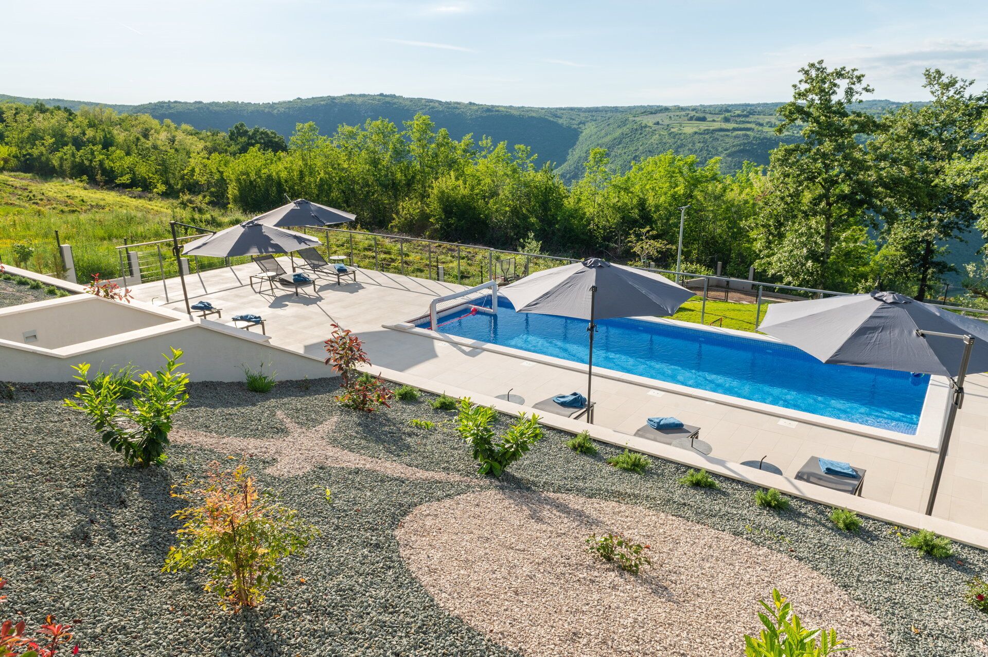 Una piscina con lettini e ombrelloni, circondata da un'area verde con piccole piante e vista su colline lontane.