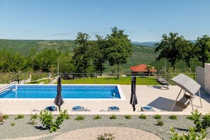 Une piscine entourée de chaises longues et de parasols, elle-même bordée d’un espace paysager offrant une vue sur un bâtiment aux toits rouges et sur des collines en arrière-plan.