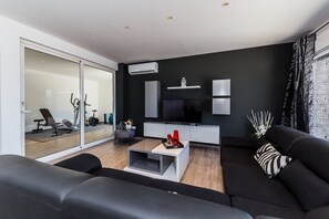 Uma sala de estar moderna com um sofá preto, uma mesa de centro branca e uma TV. Há uma porta de vidro deslizante que dá para uma área de ginástica.