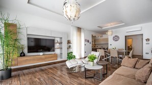 Ett modernt vardagsrum med en stor platt-TV, bekväma sittplatser, en matplats och ett kök i bakgrunden.