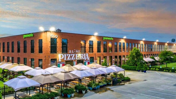 Un bâtiment dont la façade porte une enseigne indiquant « Greenworks » et le chiffre « 500 », et où se trouve au rez-de-chaussée une pizzeria appelée « Alino Pizzeria ». Devant le bâtiment, on trouve des parasols et des plantes en pot.